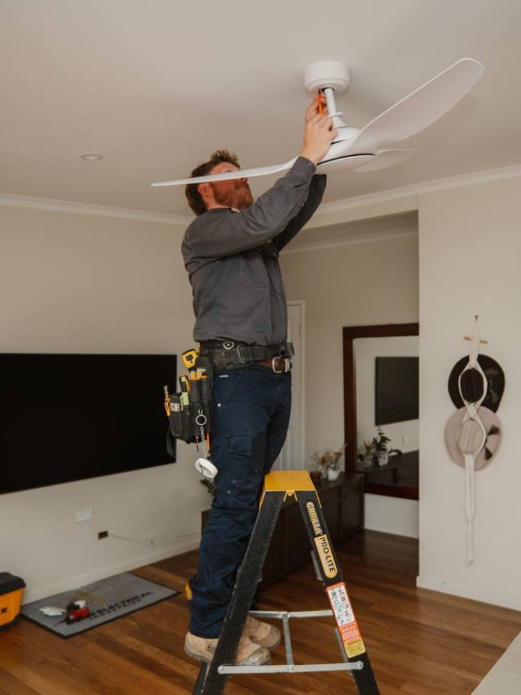 Egan Electrical electrician on a ladder installing a white ceiling fan in a living room